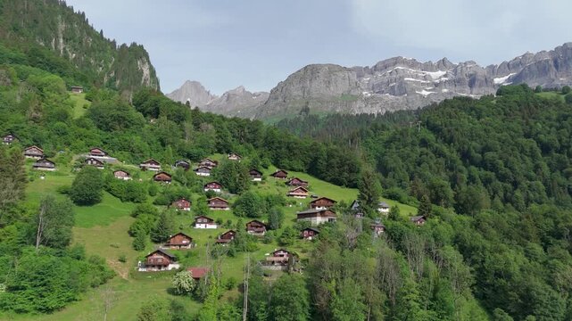 Rows of traditional alpine chalets dot lush terraced hillside beneath dramatic rocky peaks and remnant glaciers on summer day in Swiss Alps, Canton Glarus, Switzerland. Aerial drone approach