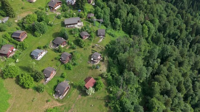 Scattered alpine chalets and cottages nestle among lush green meadows, garden plots and dense forest canopy on sunny hillside in Luchsingen, Canton Glarus, Switzerland. Aerial drone top down rise