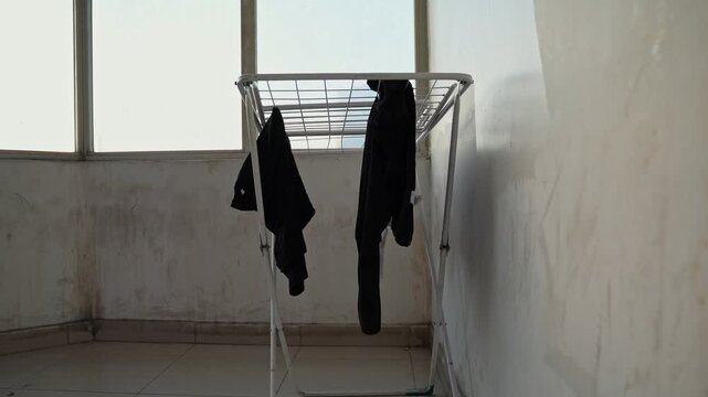 Black clothes hanging on a metal laundry drying rack inside a dirty, empty room. Bright daylight shines through large windows onto the tiled floor in this sparse, slightly rundown indoor setting.