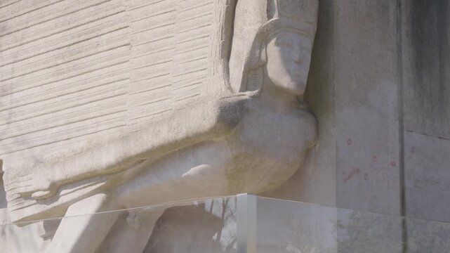 Close view of Oscar Wilde's tomb at Pere Lachaise Cemetery in Paris, showing Jacob Epstein's winged stone sculpture behind the protective glass barrier in daylight.