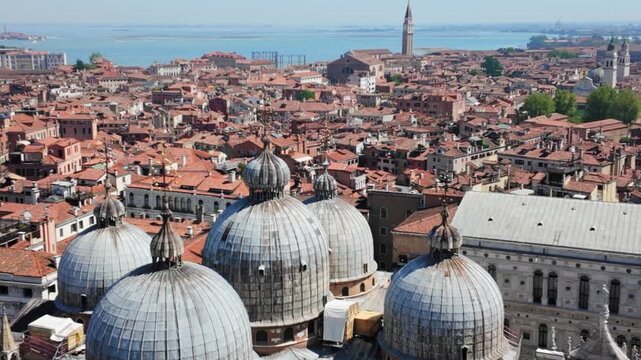 Aerial panning time-lapse from left to right over the historic lead domes of St. Mark's Basilica and traditional terracotta rooftops on a sunny day in Venice, Italy.