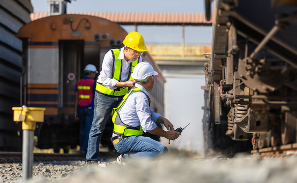 Asian railway inspector checking freight train suspension and wheel assembly at railyard, Team of Engineers in hardhat and safety vests performing maintenance inspection on cargo train equipment