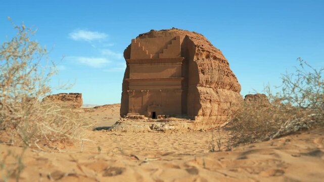 Tomb Lihyan Son of Kuza at Hegra, Saudia Arabia, low ground view, dry shrubs moving in calm wind near, sandy desert landscape around
