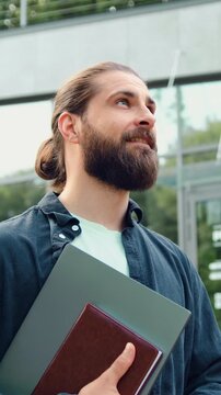Side view of young man in casual clothes is confidently walking through city park while holding laptop, with green trees and modern glass buildings in the background