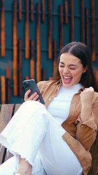 Smiling woman checking her smartphone and reacting to happy news outdoors, with a modern office building in the background. Slow motion