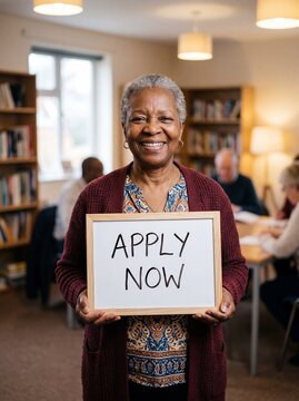 Smiling senior African American woman holding an Apply Now sign in a community library