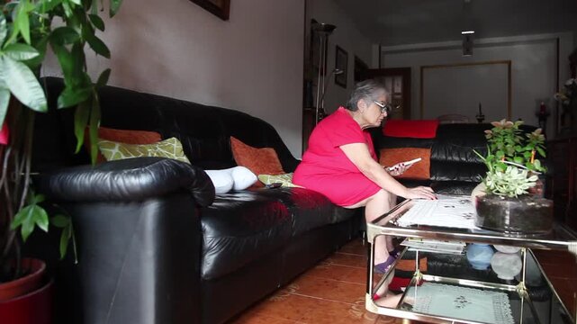  Mujer mayor con bata rosa usando un tel&eacute;fono inteligente en el sof&aacute; de su casa. Mujer mayor con gafas navegando por el tel&eacute;fono inteligente en casa. Senior activo, estilo de vida tecnol&oacute;gico moderno.