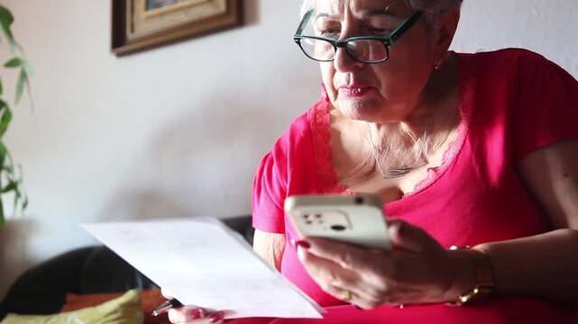  Mujer mayor con bata rosa usando un tel&eacute;fono inteligente en el sof&aacute; de su casa. Mujer mayor con gafas navegando por el tel&eacute;fono inteligente en casa. Senior activo, estilo de vida tecnol&oacute;gico moderno.