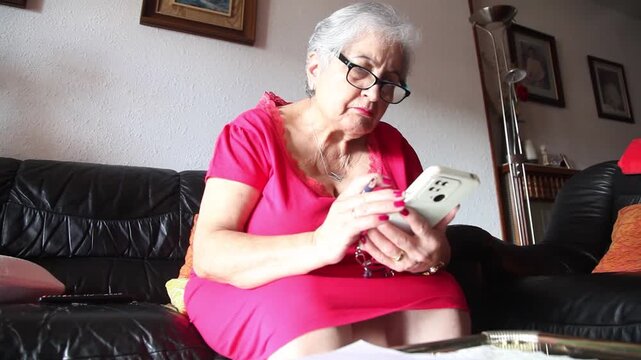  Mujer mayor con bata rosa usando un tel&eacute;fono inteligente en el sof&aacute; de su casa. Mujer mayor con gafas navegando por el tel&eacute;fono inteligente en casa. Senior activo, estilo de vida tecnol&oacute;gico moderno.