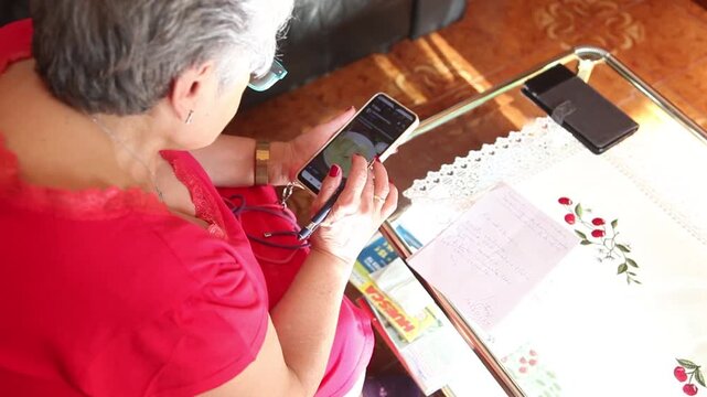  Mujer mayor con bata rosa usando un tel&eacute;fono inteligente en el sof&aacute; de su casa. Mujer mayor con gafas navegando por el tel&eacute;fono inteligente en casa. Senior activo, estilo de vida tecnol&oacute;gico moderno.