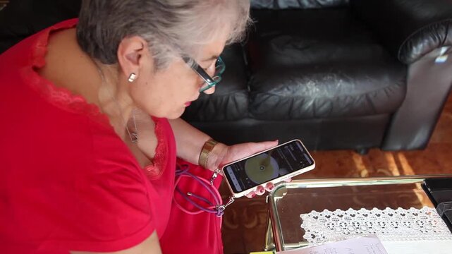  Mujer mayor con bata rosa usando un tel&eacute;fono inteligente en el sof&aacute; de su casa. Mujer mayor con gafas navegando por el tel&eacute;fono inteligente en casa. Senior activo, estilo de vida tecnol&oacute;gico moderno.