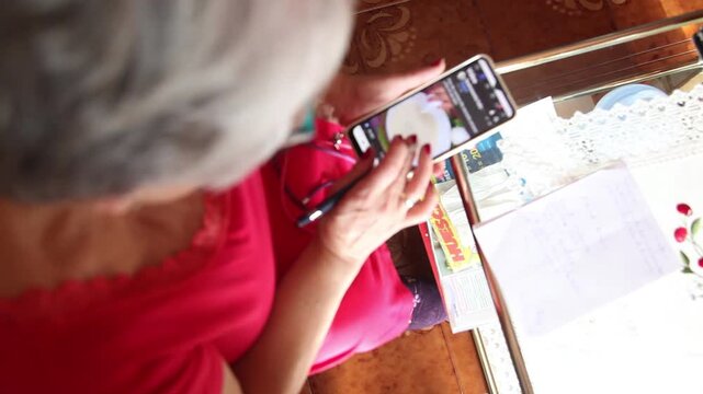  Mujer mayor con bata rosa usando un tel&eacute;fono inteligente en el sof&aacute; de su casa. Mujer mayor con gafas navegando por el tel&eacute;fono inteligente en casa. Senior activo, estilo de vida tecnol&oacute;gico moderno.