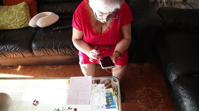  Mujer mayor con bata rosa usando un tel&eacute;fono inteligente en el sof&aacute; de su casa. Mujer mayor con gafas navegando por el tel&eacute;fono inteligente en casa. Senior activo, estilo de vida tecnol&oacute;gico moderno.
