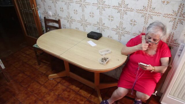  Mujer mayor con bata rosa usando un tel&eacute;fono inteligente en la silla de la cocina. Mujer mayor con bata rosa y gafas navegando por un tel&eacute;fono inteligente sentada en la cocina. Senior activo, rutina 