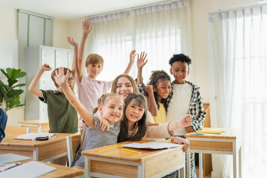 Diverse group of happy primary school children raising hands in classroom, Cheerful multi-ethnic students and teacher having fun in a modern classroom, Education concepts