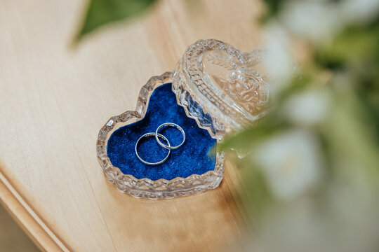 Two wedding rings resting on blue velvet inside a crystal heart-shaped box on a wooden surface. Copy space