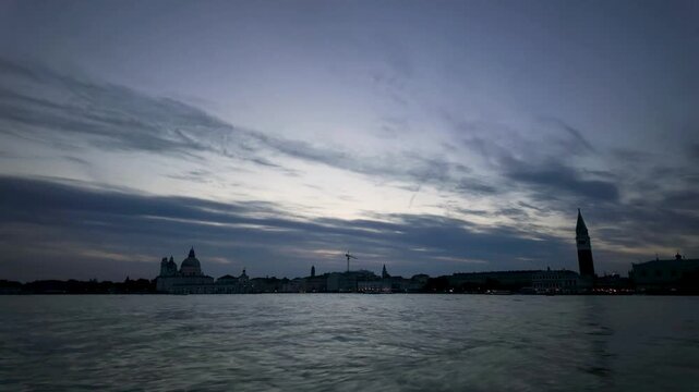 Boats navigate the Venetian lagoon during a panning day-to-night time-lapse. City lights illuminate the historic skyline featuring St. Mark's Campanile in Venice, Italy.