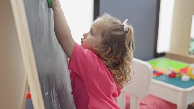 Girl with blonde curls holds a green eraser and erases a chalkboard while seated on a small stool in a playroom building; childhood curiosity.