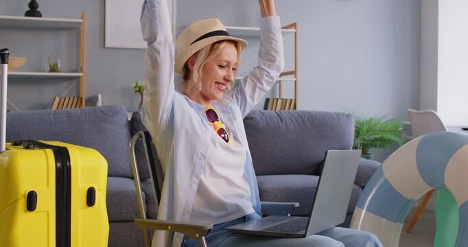 Happy excited young woman sitting at home and booking tickets online via laptop. Smiling girl in beach hat choosing tour for holiday trip. Vacation concept. 4k video. Slow motion video.