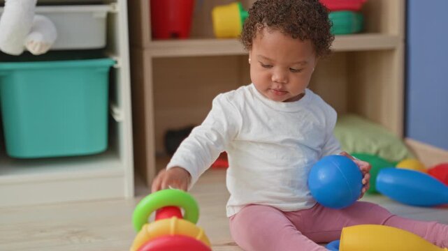 Little girl holding a blue ball reaches with her hand toward colorful stacking rings on the floor in a bright playroom with storage bins; curiosity exploration.