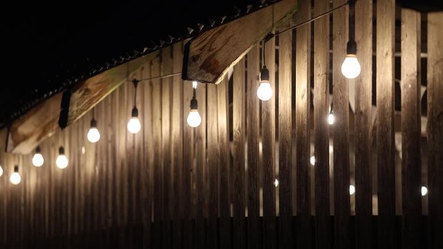 Glowing string light bulbs suspended against a rustic wooden slat wall at night, creating a warm, cozy ambiance with shallow depth of field and soft bokeh