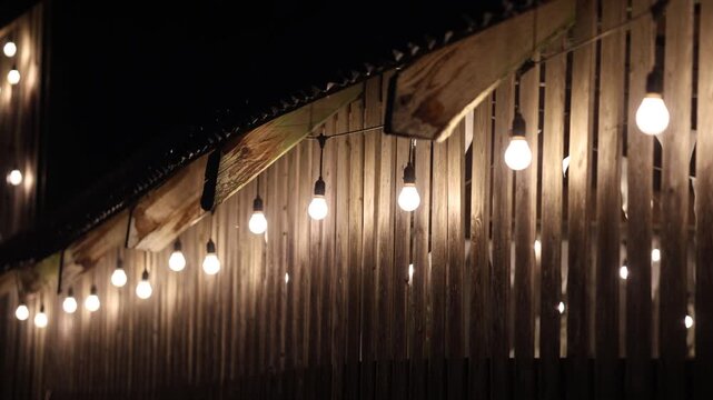 Glowing string light bulbs suspended against a rustic wooden slat wall at night, creating a warm, cozy ambiance with shallow depth of field and soft bokeh