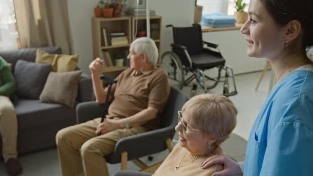 Side view shot of senior woman smiling while young female nurse in scrubs stands beside her in nursing home common room. Ideal for senior care and assisted living facility ads