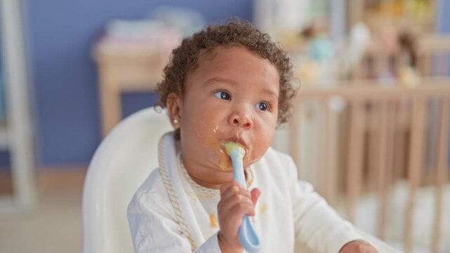 Little girl holding spoon to mouth with messy bib and food on face in building highchair; playful curiosity.