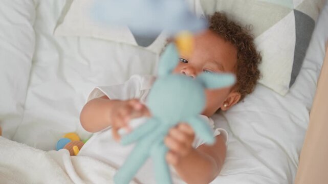 Little girl reaching and holding a blue stuffed rabbit with outstretched hand on a bed in building; tender curiosity.