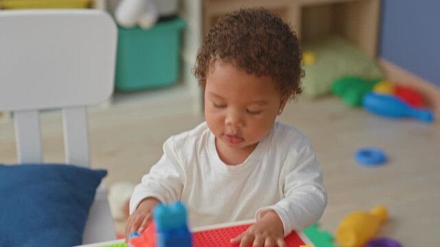 Little african american girl plays with plastic building blocks, placing studs on a red baseplate with her hands at a home playroom table; curiosity.