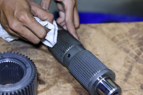 Close up view of a technician's hands using a white cloth rag to clean a metallic transmission splined shaft component at a vehicle repair workshop.