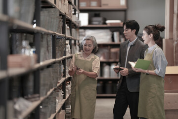 Fototapeta premium A young Asian craft shop owner in a product storeroom discussing inventory together in a dark wood-shelved storeroom in a small business warehouse. A picture of small business success.