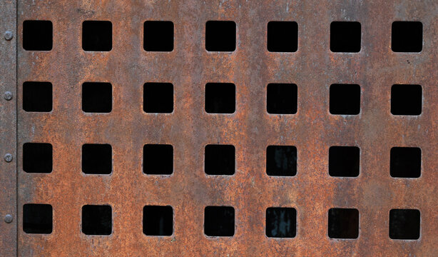 Close up of rusted metal surface with pitted corrosion, 7x4 cutout grid pattern, dark openings, and vertical row of rivets on the left under even lighting