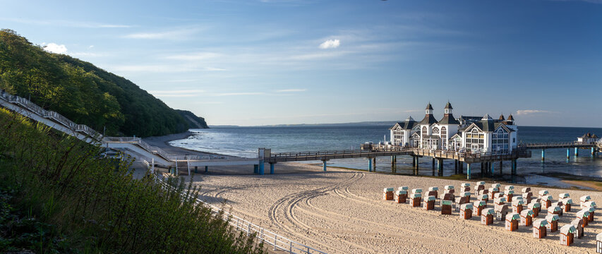 Panoramic landscape view of the Baltic Sea coastline in Sellin, featuring the famous wooden pier, sandy beach, traditional beach chairs, and a large wooden staircase on Ruegen, Germany.