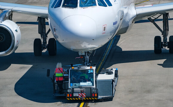 Direct Frontal Perspective Of A Large Commercial Passenger Airplane Being Carefully Guided And Pushed By A Specialized Airport Ground Support Tractor On The Tarmac