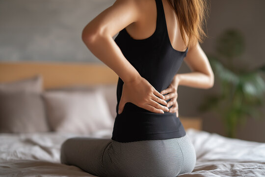  A woman sits on her bed holding her lower back with both hands, signaling discomfort and muscle pain.
