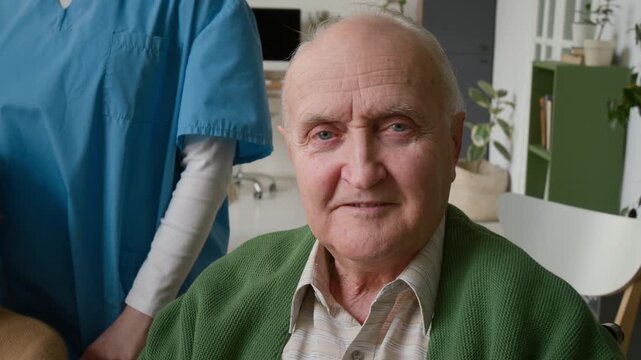 Portrait of senior man looking at camera and smiling in retirement home with caregiver in blue uniform standing nearby. Video promoting assisted living and senior care