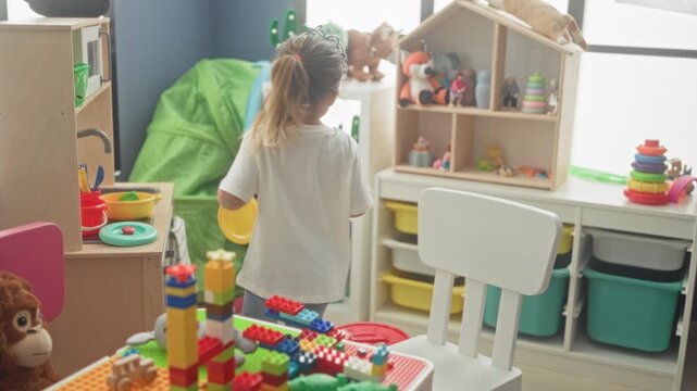 Girl wearing tiara holds toy discs by shelving and chair in a playroom inside a building; thoughtful play.