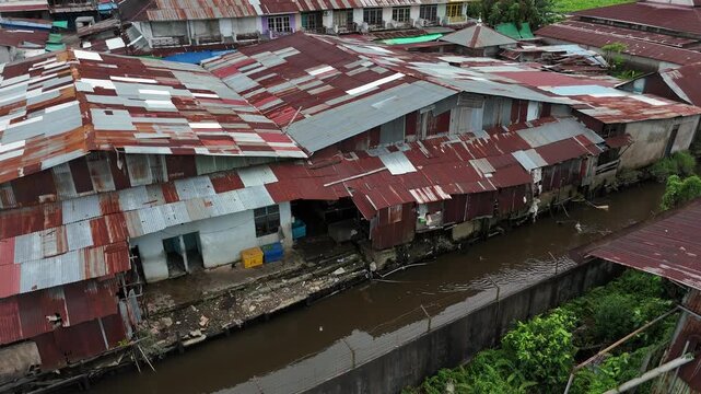 An aerial view showing a poor riverside shantytown with rows of rusty corrugated tin roofs lining a muddy, brown canal in Indonesia.