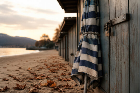 Beach huts lined along the shore with a striped umbrella leaning against one. Tranquil seaside atmosphere during sunset.