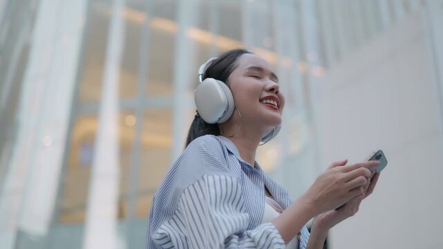 Happy Asian woman listening to music with headphones and holding smartphone, representing city lifestyle professional women relaxing, positive emotion energy and audio streaming app