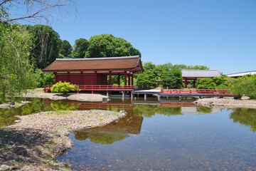 Fototapeta premium Nara Palace Site reflecting traditional Japanese architecture. Traditional red palace buildings are reflecting in a calm pond surrounded by green trees and a blue sky.