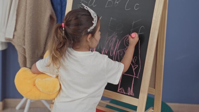 Little girl drawing on chalkboard with pink chalk, wearing tiara and plush duck backpack in classroom; playful creativity.