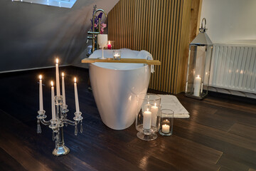 Luksusowy salon kąpielowy na poddaszu z wolnostojącą wanną, urządzony w przytulnym, eleganckim stylu z drewnianymi belkami, nastrojowymi świecami i kolorowym fotelem akcentującym. © arteffect.pl