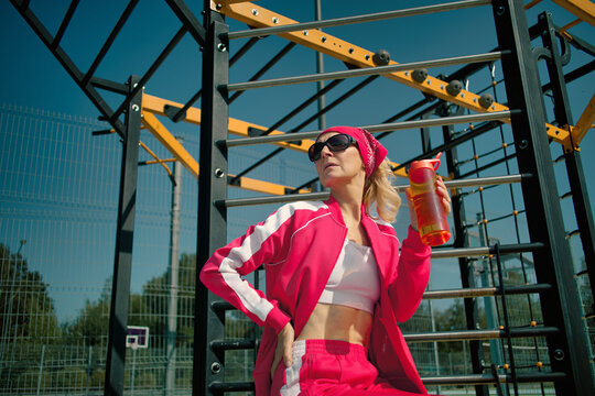 Active 60 years old senior woman in pink tracksuit and sunglasses holding water bottle at workout park