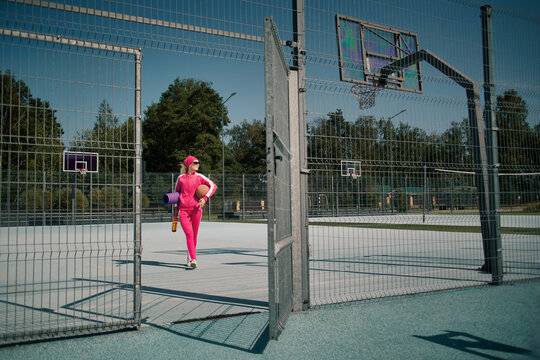 Active 60 years old senior woman in pink tracksuit holding sports equipment walking through open court gate