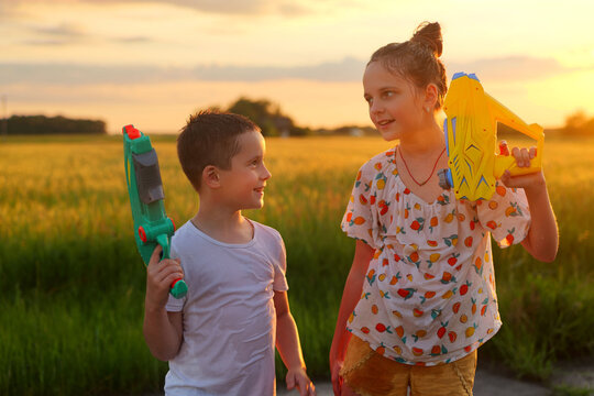 Two children are having fun with water guns in a green field at sunset. They look happy while enjoying their time outside in warm weather during summer