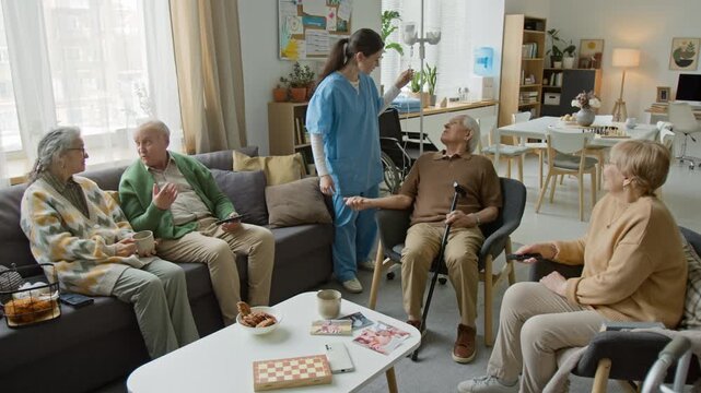 Wide angle shot of female nurse in scrubs talking to seniors in retirement home common room. Suitable for senior care facility and medical insurance advertising