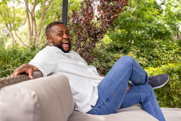 Obraz premium Adult African man reclining on cushioned wicker sofa wearing white shirt and blue jeans on patio