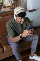 Obraz premium African American male sitting at home on brown sofa, wearing headphones and holding blue smartphone